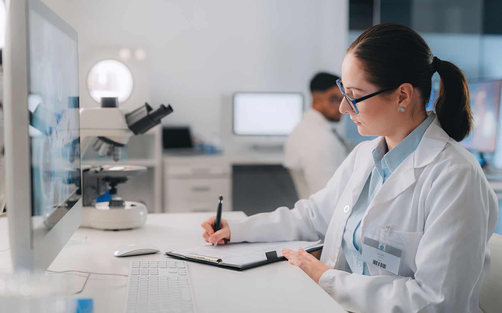 woman scientist doing research and writing in laboratory for experiment. analyze results on computer, microscope and clinical diagnosis notes, pharmacy and lab technician or medical assistant.
